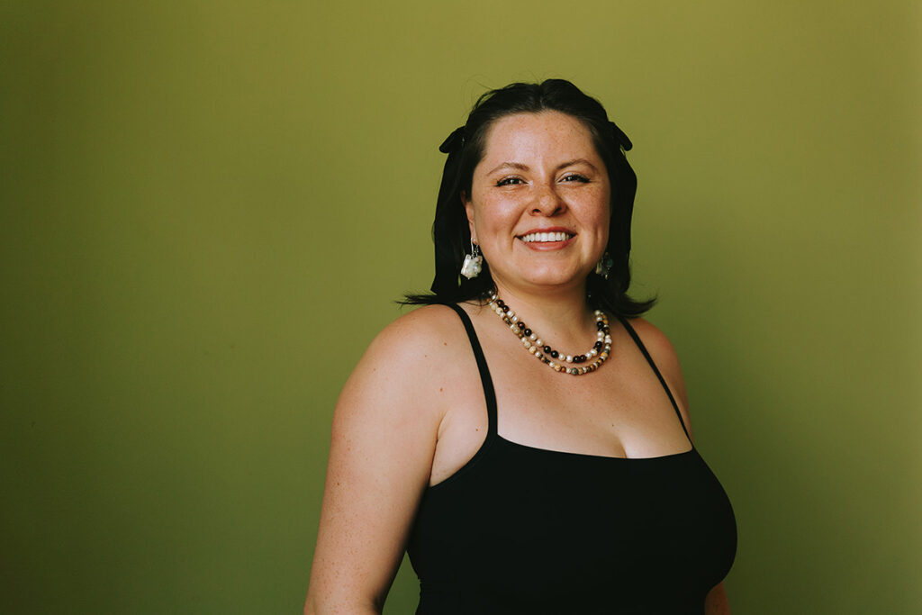 Headshot of Paige Brown, the artist, smiling at camera, wearing pearl earrings and necklaces, with a light mossy green background. 